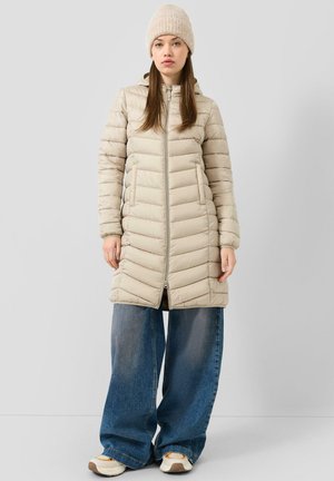 Jeune femme portant un manteau beige matelassé à capuche, un bonnet en tricot, un jean bleu à jambes larges et des baskets blanches, debout devant un fond uni.