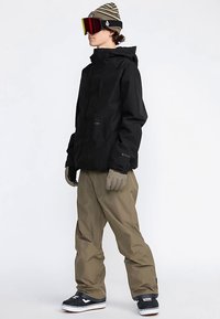 Black waterproof jacket with a hood, paired with beige snow pants. Accessories include gloves and a beanie with stripes. Snowboard shoes.