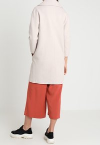 Světle růžový kabát s klopovým límcem, nošený přes oranžové kalhotové sukně. Černé tenisky doplňují outfit. Hladká textura, střih na míru.