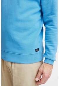 Sudadera azul claro con dobladillo y puños acanalados. Presenta una pequeña etiqueta negra en el lado inferior. Textura suave, cuello redondo y ajuste casual.