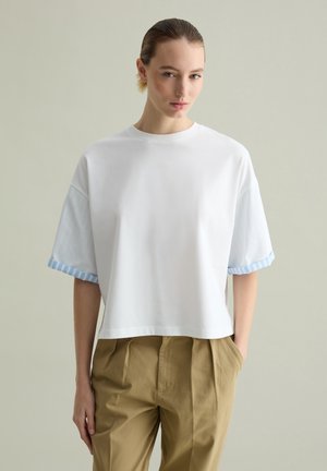 White cropped t-shirt with short, light blue striped sleeves. Smooth fabric, relaxed fit, minimalistic design, paired with beige trousers.