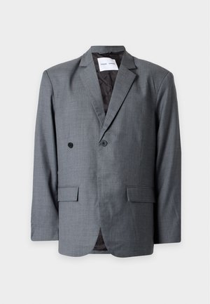 Homme debout portant un blazer gris à double boutonnage, une chemise blanche boutonnée, un pantalon noir à jambes larges et des chaussures noires sur un fond uni.