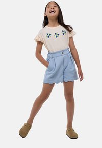 T-shirt crème à manches courtes avec broderie florale, assorti à un short bleu clair à bords festonnés. Le modèle porte des sabots marron.