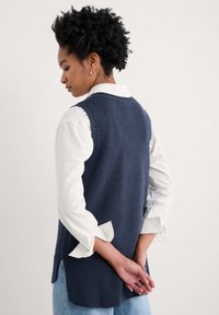 Chaleco sin mangas de punto en color navy con dobladillo acanalado, estilizado sobre una camisa blanca de botones, que presenta un cuello redondo clásico y aberturas laterales.