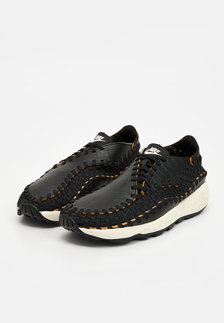 Baskets tissées noires avec un matériau texturé et des accents de couture orange, et une semelle blanche. Conçues pour le confort et la flexibilité.
