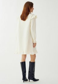 Valge tekstuuriga kleit volangidega õlgadel ja hemmi ääres, koos mustade läikivate põlvekõrguste saapadega. Puhtad, minimalistlikud jooned.