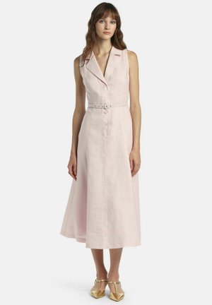 Frau trägt ein ärmelloses, blassrosa Midi-Kleid mit Knöpfen, einem passenden Gürtel und goldfarbenen, spitz zulaufenden T-Riemchen-Schuhen, steht vor weißem Hintergrund.