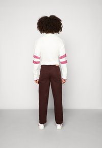 Persona con cabello rizado lleva un suéter blanco con rayas rosas en las mangas, pantalones marrones y zapatillas blancas, de pie frente a una pared lisa.