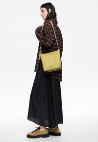 Borsa a tracolla gialla in pelle con forma piatta, dotata di una base strutturata e accents marroni. Indossata con una camicia a quadri e una gonna nera.
