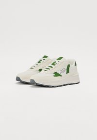 Vajszínű anyagú felnőtt sneakers, zöld részletekkel és fehér fűzővel. A talp felszíne strukturált, ami biztosítja a tapadást. Az oldalán található címke tartalmazza a márkainformációt.