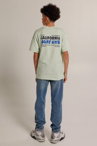 T-shirt in cotone verde menta con una vivace stampa grafica di testo in blu e nero sul retro, abbinato a jeans in denim azzurro e scarpe sportive.