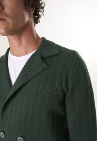 Giacca in maglia verde a doppio petto con colletto ampio e motivo esagonale. Presenta grandi bottoni e una finitura morbida e tessuta.
