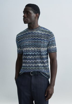 Kortärmad skjorta med flerfärgat zickzackmönster i nyanser av blått, grönt och grått. Bärs med mörka byxor med mönster och ett bälte.