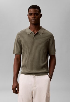 Homme portant un polo côtelé vert olive à manches courtes et un pantalon beige clair, debout avec une main dans la poche sur un fond uni.