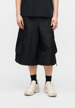 Person iført sorte cargoshorts, sorte sokker, beige sneakers og en sort skjorte med et lille mærke nær kanten.
