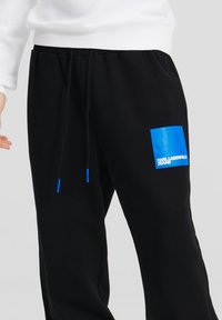 Schwarze Jogginghose mit elastischem Bund, mit einem blauen quadratischen Akzent am Oberschenkel und blauen Kordelzügen. Text: "Karl Lagerfeld Jeans."