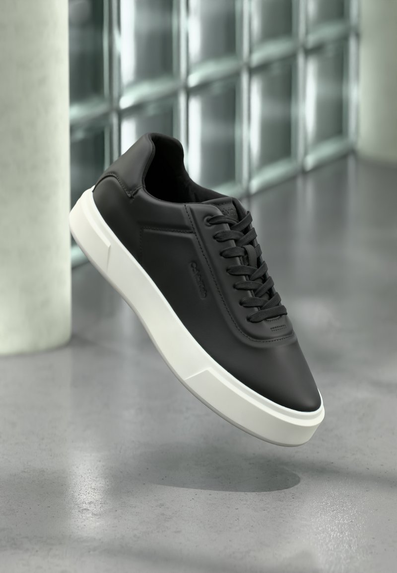 Schwarze Ledersneaker mit weißer Gummisohle, rundlicher Zehenpartie und flachen Schnürsenkeln. Strukturierte Akzente an den Seiten, minimalistisches Design.