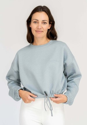Frau mit braunen Haaren richtet hellblauen Sweatshirt mit Kordelzug, trägt weiße Hose und steht vor neutralem Hintergrund.