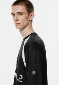 Schwarzes Langarmshirt mit weißen Akzenten und einem kreisförmigen Logo mit Spielcontroller-Tasten auf dem linken Ärmel. Weicher Stoff, entspannte Passform.