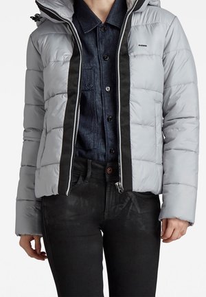 Personne portant une doudoune gris clair par-dessus une chemise à boutons bleu foncé et un pantalon noir, montrée du cou aux hanches.