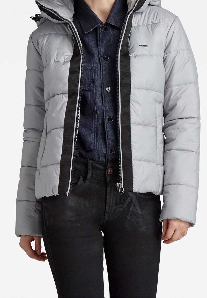 Personne portant une doudoune gris clair par-dessus une chemise à boutons bleu foncé et un pantalon noir, montrée du cou aux hanches.