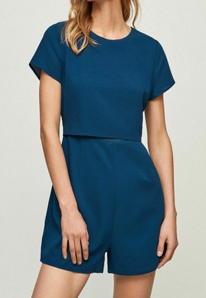 Woman wearing a dark teal short-sleeve romper with a layered crop top and high-waist shorts against a neutral background.