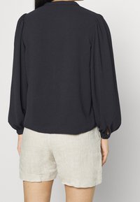 Blouse marine à manches bouffantes avec une finition texturée, présentant un ourlet arrondi et des ouvertures discrètes aux poignets des manches, portée avec un short beige clair.