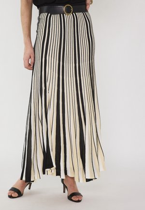 Jupe longue à rayures verticales noires et blanches avec des plis, assortie à une ceinture noire avec une boucle circulaire dorée et des talons ouverts noirs.