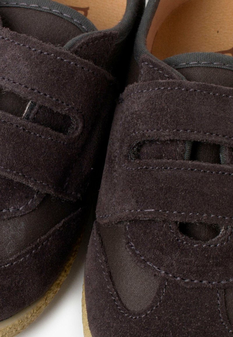 Pantofi din suede maro închis, cu două curele orizontale Velcro, margini cusute și o talpă din cauciuc texturat. Au vârf rotunjit și accente decupate.