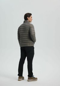 Polstret grå jakke med høj krave, letvægtsdesign og vandret syning, båret med sorte bukser og sandfarvede vandresko.