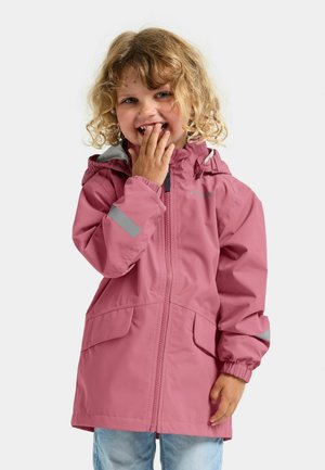 Krulligharig kind in een roze regenjas met reflecterende patches, glimlachend en met één hand de mond bedekkend, staand tegen een witte achtergrond.