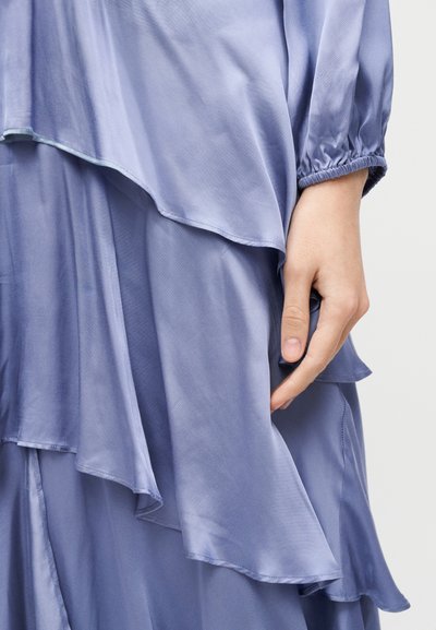 Το χέρι μιας γυναίκας ακουμπάει δίπλα σε αέρινο φόρεμα από στρωματοποιημένο ανοιχτό μοβ σατέν με ελαστική μανσέτα μακριού μανικιού.