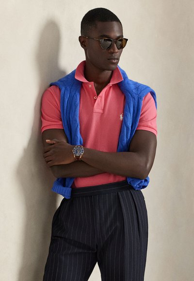 Uomo con occhiali da sole tartarugati, polo rosa con maglione blu sulle spalle, pantaloni scuri a righe e orologio da polso, braccia incrociate.