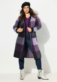 Langer Cardigan in Violetttönen mit einem karierten Muster, kombiniert mit einem lila Top und dunklen Jeans. Silberne Knöchelstiefel runden den Look ab.