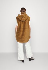 Brauner, ärmelloser Hoodie-Westen mit sanfter, flauschiger Textur, abgerundetem Saum und großer Kapuze. Über einem weißen Oberteil getragen und mit weißen Hosen kombiniert.