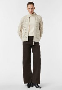 Kobieta stoi w beżowym swetrze ze splotem, pod którym ma kremowy golf, ciemnych szerokich spodniach i czarnych botkach za kostkę na białym tle.