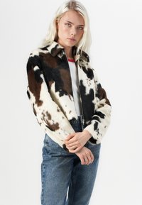 Jachetă scurtă din piele de vacă maro și alb, cu o textură pufoasă, fermoar frontal și guler. Asortată cu blugi din denim albastri și un top alb.