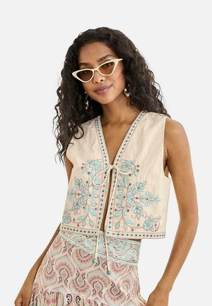 Mujer con cabello largo y rizado que lleva un chaleco sin mangas color crema con bordado floral azul, gafas de sol tipo ojos de gato y falda con estampado y cintura de encaje.