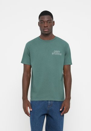 T-shirt verde a maniche corte in cotone. Presenta un colletto girocollo a costine e un piccolo logo "OBEY STUDIOS" bianco sul petto.