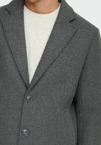 Grau melierter Woll-Mix-Mantel mit Reverskragen, zwei Knöpfen und Seitentaschen. Über einem hellen Strickpullover getragen. Strukturierten Stoff.