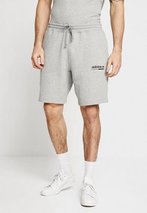 Homme portant un short Adidas gris clair à cordon, des chaussettes blanches et des baskets blanches, debout sur un fond blanc uni.