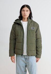 Olivžalių spalvos paminkštintas džemperis su užtrauktuku, aukšta apykakle ir siuvinėtu logotipu ant krūtin ės, derinamas su šviesiai mėlynais džinsais.