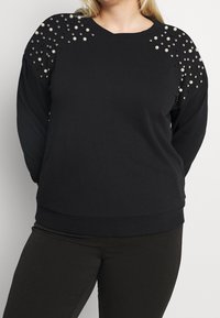 Svart sweatshirt med långa ärmar, prydd med pärlor och metallnitar på axlarna, med rundad halsringning och ribbat avslut.
