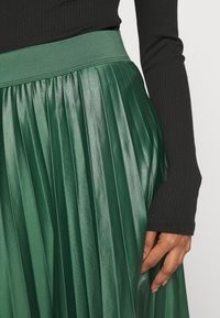 Žena v lesklé plisované zelené sukni a černém žebrovaném topu s dlouhým rukávem, s pravou rukou odpočívající na sukni.