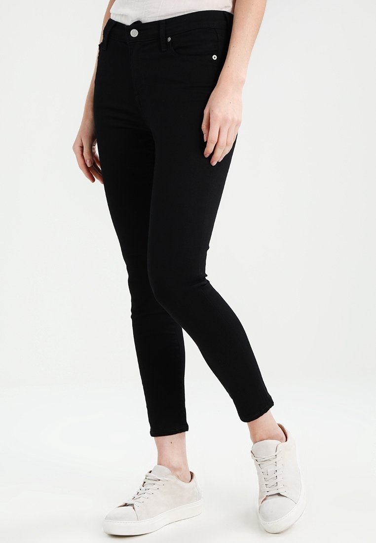 Jeans skinny de mezclilla negra con cinco bolsillos, cierre frontal de botón y un ajuste ceñido. Combinados con zapatillas de color claro.