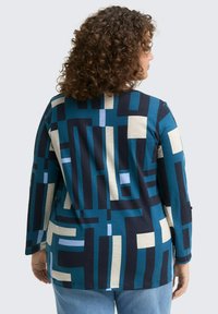 Vrouw met krullend haar die een langarmige geometrisch patroonshirt in teal, marineblauw, crème en lichtblauw draagt, met de rug naar de camera tegen een effen achtergrond.