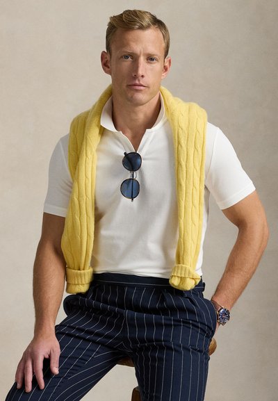 Geltonas megztinis perrištas per baltą polo marškinėlį, derinamas su tamsiai mėlynomis dryžuotomis kelnėmis. Aksesuarai: apvalios saulės akiniai ir laikrodis.