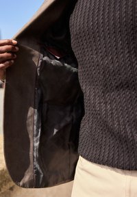 Giacca in suede marrone aperta per rivelare un interno nero liscio con un motivo a diamante e dettagli in bordeaux. Maglione scuro con texture visibile sotto.