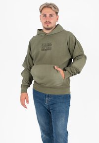 Olijfgroene hoodie met oversized ontwerp, voorste kangoeroezak en een merklogo. Gecombineerd met blauwe jeans staat het model tegen een witte achtergrond.