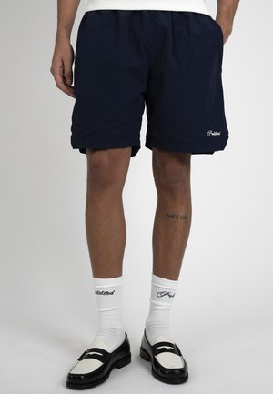 Uomo che indossa pantaloncini blu navy, calzini bianchi con scritte nere e mocassini bianco-neri, in piedi su uno sfondo semplice.